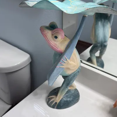 Hand-carved and painted wooden frog figurine holding a lotus leaf umbrella, featuring whimsical details and a pastel color palette, ideal...