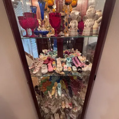 Vintage wooden corner curio cabinet filled with collectible glassware and miniature shoes, featuring Fenton hobnail glass goblets, amber...