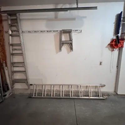 Three aluminum ladders, including a tall extension ladder, a medium step ladder, and a small step stool, stored in a garage setting.