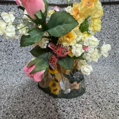 Decorative faux floral arrangement featuring pink roses, yellow blossoms, and a butterfly accent, housed in a garden-themed resin base...