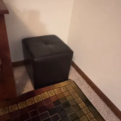 Contemporary dark brown faux leather cube ottoman with a single tufted button on top, positioned in a room corner.