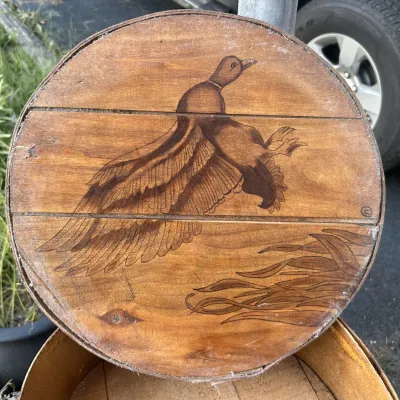Vintage round wooden cheese box lid featuring a pyrography design of a mallard duck in flight over reeds.