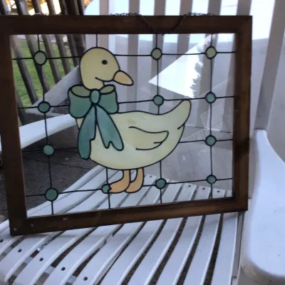 Vintage stained glass window panel featuring a yellow duck wearing a blue bow, set in a dark wood frame with a hanging chain.