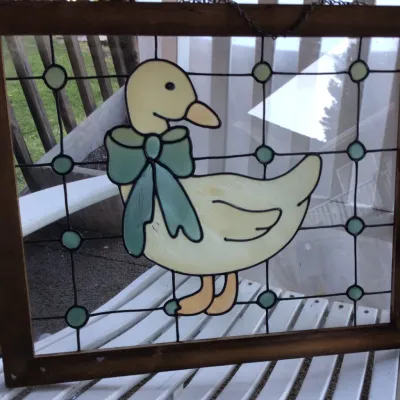 Vintage country-style stained glass window panel featuring a yellow duck with a green bow, housed in a simple wooden frame with a hanging...