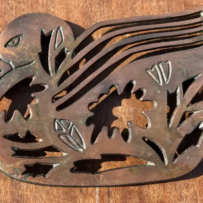 Vintage cast brass or bronze trivet shaped like a duck, featuring stylized cutouts of flowers and foliage with a natural aged patina.