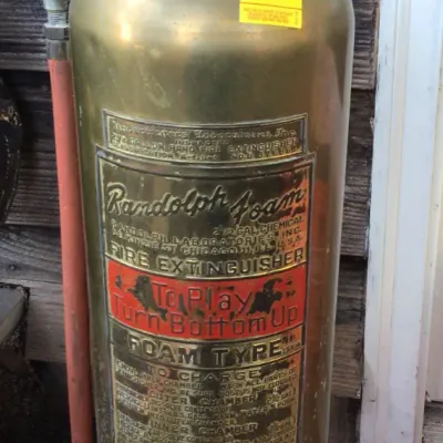 Vintage Randolph Foam 2.5-gallon brass fire extinguisher with red painted top handle and side hose, showing some tarnish and wear.