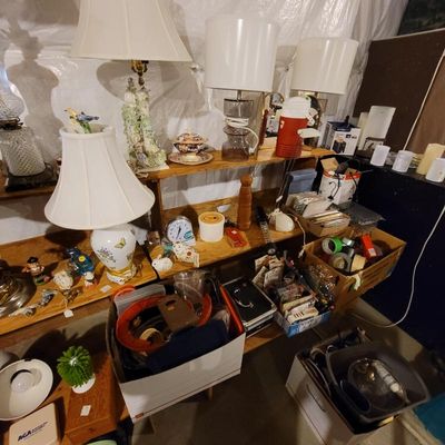 Assorted household goods, including table lamps, decorative ceramics, a small clock, and boxes of miscellaneous items on wooden shelving.