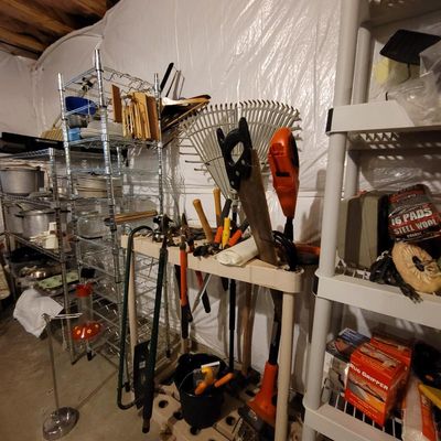 Assorted garden and hand tools, including rakes, loppers, and a hand saw, organized on shelving units alongside household items like...
