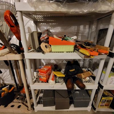 White plastic four-tier shelving unit holding various household and workshop items, including steel wool, hand tools, a DeWalt tool bag,...