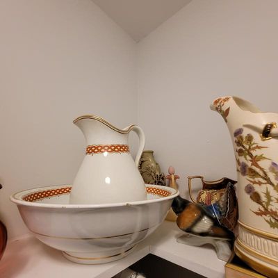 Antique ceramic wash basin and pitcher set with gold trim and a red geometric band, displayed alongside other vintage decorative pitchers...