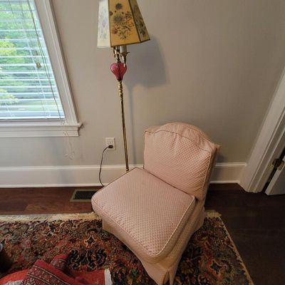 Vintage pink upholstered slipper chair with a matching skirt, paired with a brass floor lamp featuring a pink glass accent and a floral...