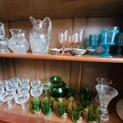 Assorted vintage glassware displayed on wooden shelves, including cut crystal pitchers, pink and blue goblets, and a green glass pitcher...