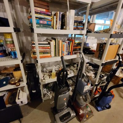 Assorted household items, including books on shelving units and several upright vacuum cleaners and carpet cleaners.
