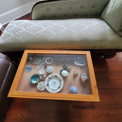 Wooden display coffee table with a glass top, showcasing various small collectibles and porcelain dishes, positioned in front of an...