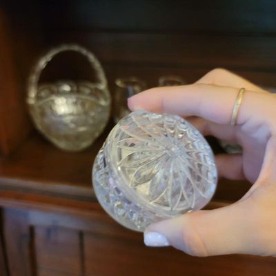 Small cut glass or crystal lidded jar with a starburst pattern on the lid, shown held in hand with other glassware in the background.