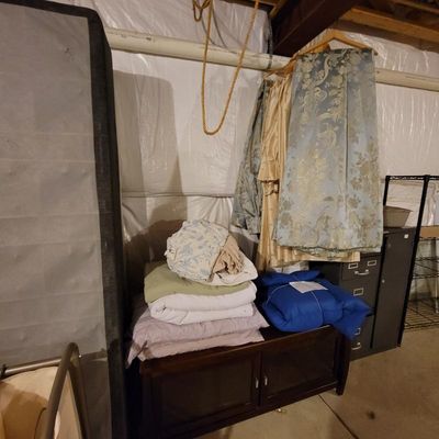Assorted household linens, including folded blankets, sheets, and hanging blue and gold damask drapery panels, displayed in a basement...