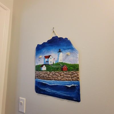 Hand-painted slate wall plaque depicting a coastal lighthouse scene, signed by the artist and ready to hang with a rope hanger.