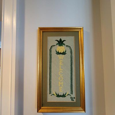 Framed needlepoint art featuring a vertical "WELCOME" design topped with a pineapple, housed in a gold-finished wooden frame.
