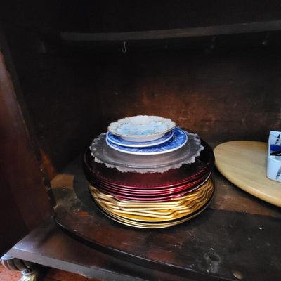 Stack of assorted decorative plates and chargers, including gold-toned, red, and blue-and-white patterned pieces, stored inside a dark...