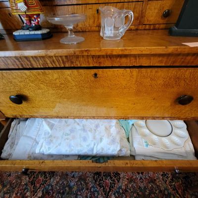 Antique bird's eye maple chest of drawers with dark wooden pulls, shown with an open drawer containing linens and a ceramic deviled...