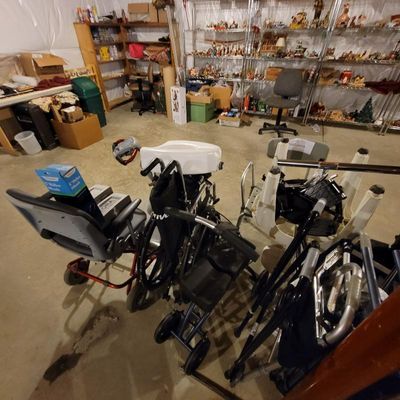 Assorted mobility aids including a wheelchair, rollator walker, and shower chairs, displayed in a basement setting with shelving in the...