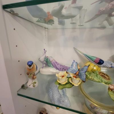 Collection of Herend porcelain figurines, including a peacock, a bluebird, and a butterfly on flowers, displayed on glass shelves.