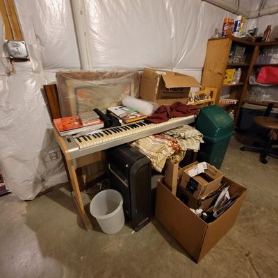 Electronic keyboard on a wooden stand, cluttered with various household items, boxes, and linens in a basement setting.