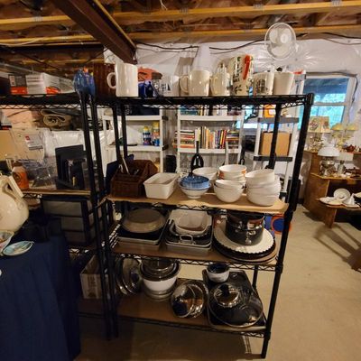 Black wire shelving unit filled with kitchenware, including ceramic mugs, mixing bowls, baking pans, and stainless steel pots with lids.