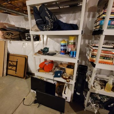 White plastic four-tier shelving unit holding various household items, including bedding, craft ribbons, and small electronics, alongside...