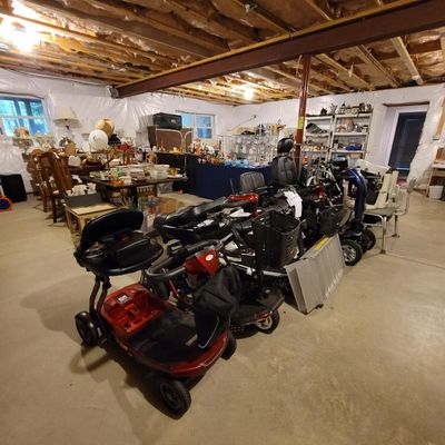 Multiple mobility scooters, wheelchairs, and a portable ramp displayed for sale in a basement alongside various household goods and...