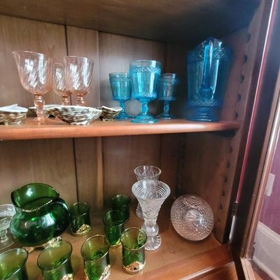 Assorted vintage glassware displayed in a wooden cabinet, featuring pink swirl goblets, blue pressed glass goblets with a matching...
