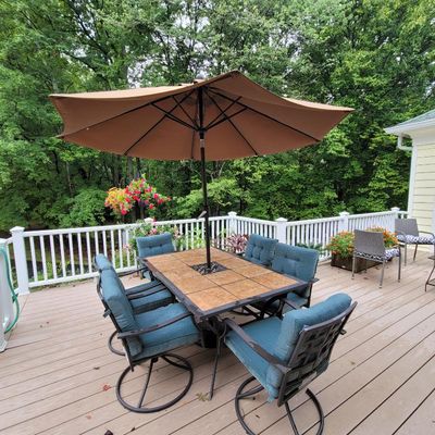 Outdoor patio dining set featuring a tile-top rectangular table, six swivel chairs with teal cushions, and a large brown market umbrella.