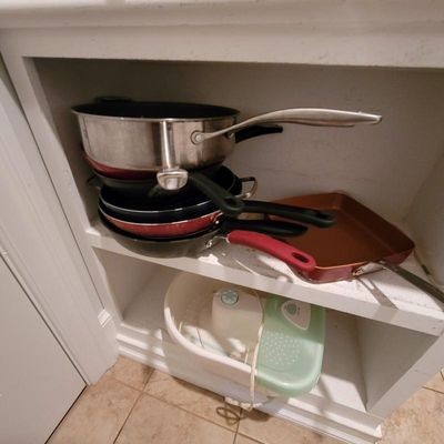 Assorted kitchen cookware, including stainless steel and non-stick frying pans, stacked on a white shelf above a vintage foot spa.