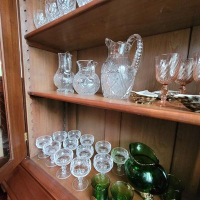 Collection of vintage glassware displayed in a wooden hutch, including cut crystal pitchers, pink depression glass goblets, and green...