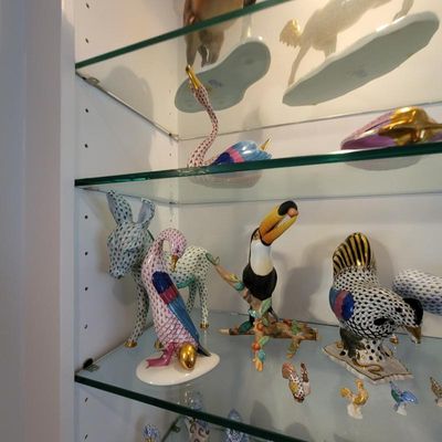 Collection of Herend porcelain animal figurines, including a toucan, donkey, and various birds, displayed on glass shelves.