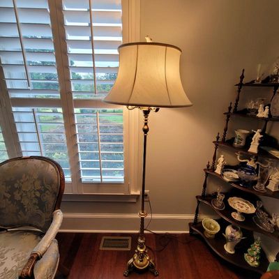 Vintage brass floor lamp with an ornate footed base and a flared fabric shade, shown illuminated in a living room setting.