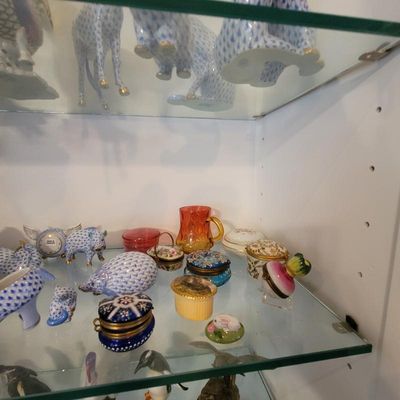 Collection of vintage porcelain trinket boxes, miniature figurines, and a small amber glass pitcher displayed on glass cabinet shelves.