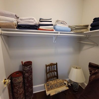Assorted bed linens stacked on a closet shelf, alongside rolled oriental-style rugs, a wooden ladder-back chair, and a ceramic table lamp.