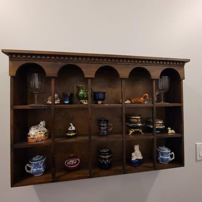 Vintage wooden wall-mounted display shelf with arched cubbies, filled with an assortment of miniature porcelain boxes, glassware, and...