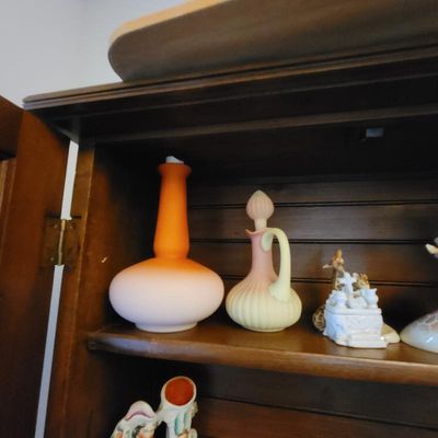 Fenton Burmese glass cruet and a satin glass peachblow vase, displayed alongside small ceramic figurines on a wooden hutch shelf.