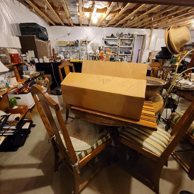 Round wooden dining table with striped upholstered chairs, shown in a basement setting with various household items and boxes.