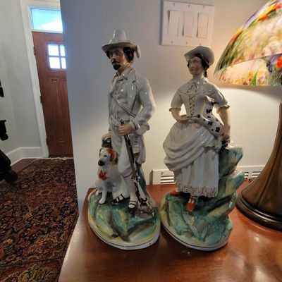 Pair of vintage Staffordshire-style porcelain figurines depicting a hunter with his dog and a woman holding a small animal, featuring...