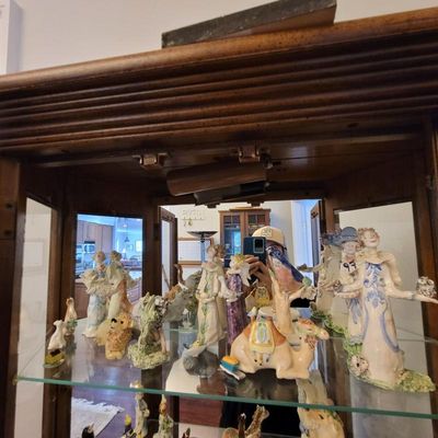 Collection of whimsical ceramic figurines, including angels, animals, and stylized people, displayed on glass shelves inside a lighted...