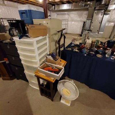 Assorted household items in a basement, including a white plastic storage tower, a Holmes floor fan, a coffee maker, and a table...