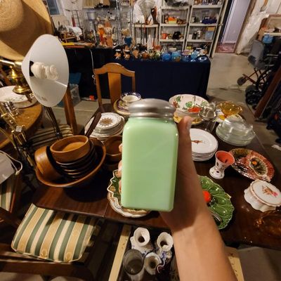 Vintage jadeite glass shaker with a metal lid, shown in hand over a table filled with various glassware, ceramics, and dining items.
