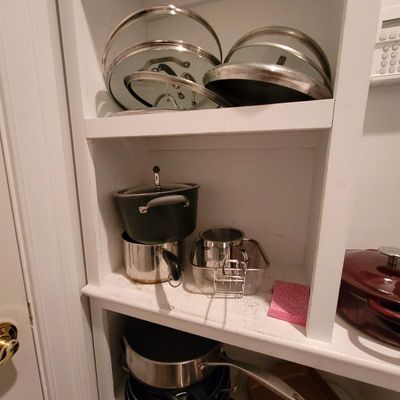 Assorted cookware, including stainless steel and non-stick pots, pans, and glass lids, stored on white built-in shelving.