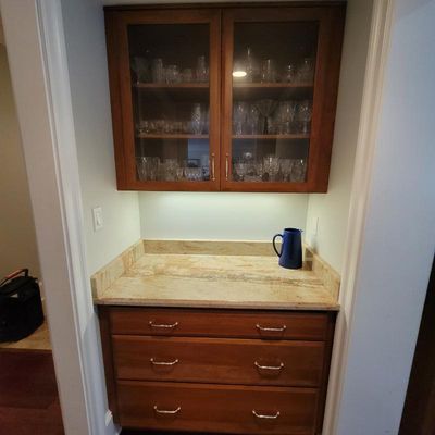 Built-in wooden bar cabinet featuring a granite countertop, three lower drawers, and an upper glass-front cabinet filled with various...