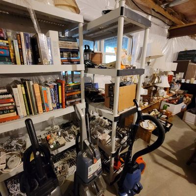 Assorted household items including books on shelving units, upright vacuum cleaners, and table lamps in a basement storage area.