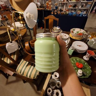 Vintage McKee jadeite ribbed shaker jar with a metal lid, held up against a background of various estate sale items.