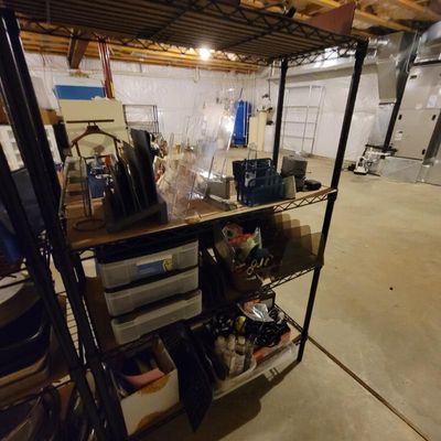 Black wire shelving unit holding various office supplies, storage bins, and household items in a basement setting.
